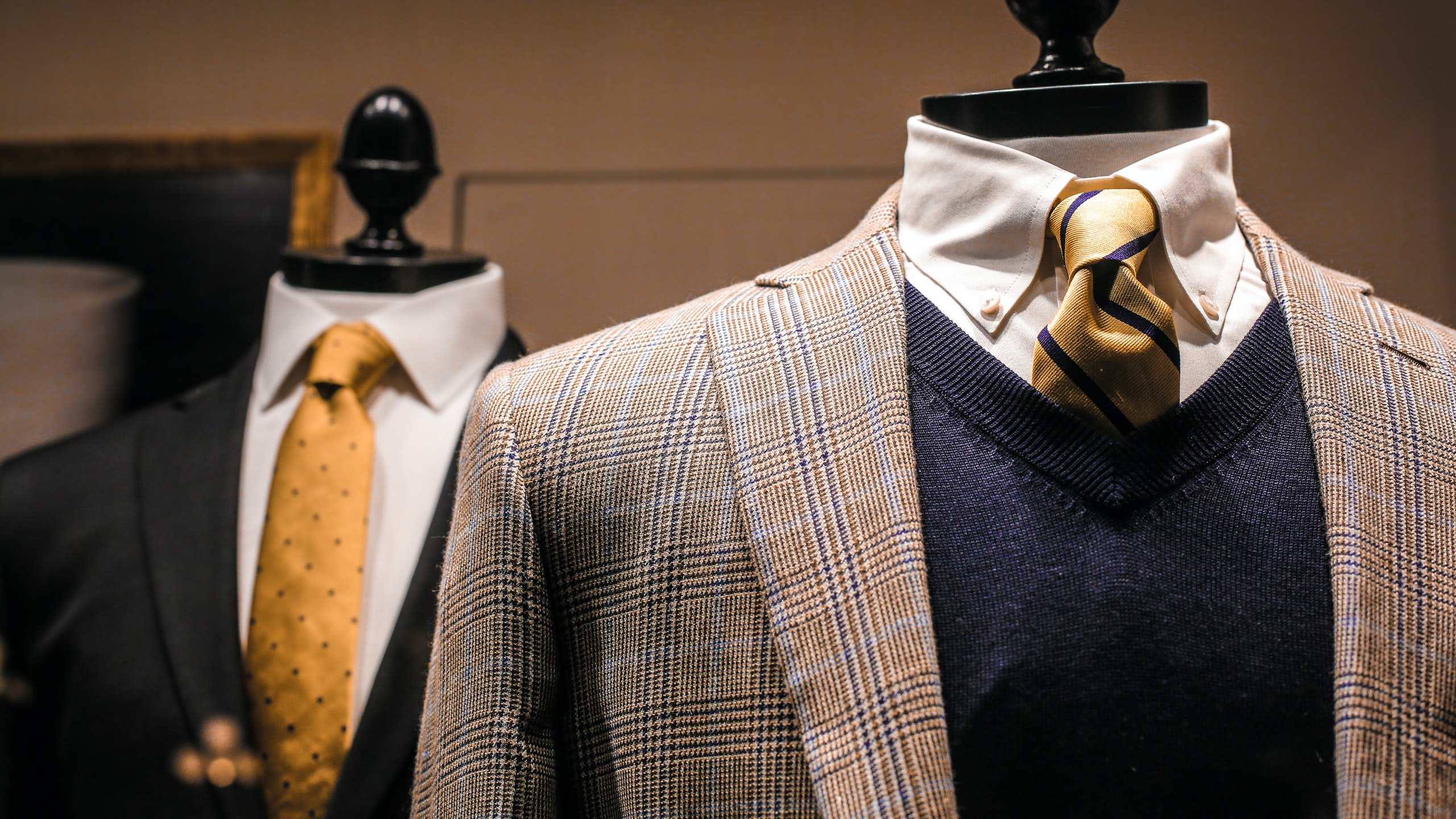 Two suits, one with beige suit and dark blue jumper. one with dark blue suit and yellow tie